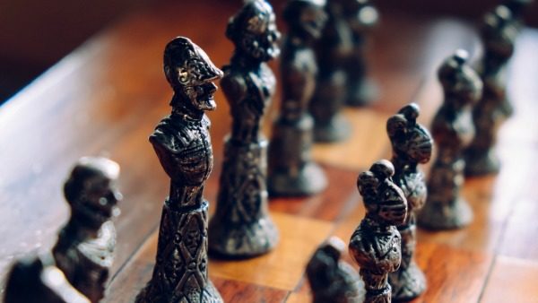 Photo credit: Lou Levit | Close up of metal chess pieces. They're arranged on a chestnut wooden chess board, facing right in relation to the camera. The metal seems to be aged.