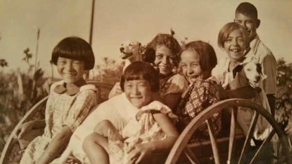 Six children—two Japanese, four White American—sit smiling in a large-wheeled wagon. The girls are wearing tunic dresses. The boy stands behind the wagon. Two girls are holding dogs in their arms.