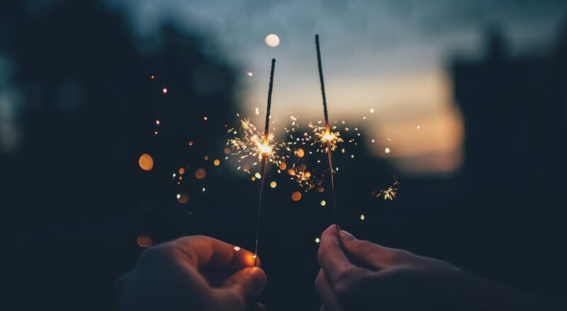 Two hands hold sparklers. Sunset in the background, out of focus.